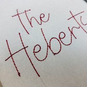 Może przedstawiać: Zbliżenie białej tkaniny wyhaftowanej słowami "The Heberts" czerwoną nicią. Pismo jest w stylu kursywy, z połączonymi literami. Tkanina ma subtelną fakturę.