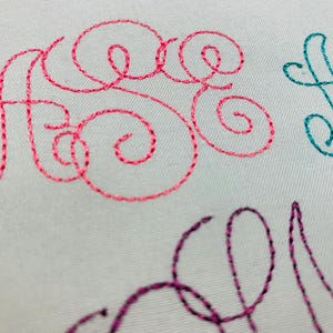 May include: Close-up of embroidered lettering on a white fabric surface. The letters are in pink, teal, and purple. The letters are in a cursive font.