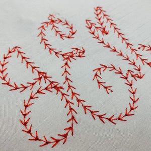 May include: A light grey fabric featuring a decorative embroidery pattern stitched in vibrant red thread. The design consists of continuous, flowing lines formed by a repeating feather stitch, creating an organic, vine-like shape across the surface.