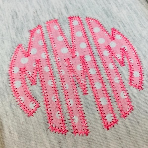 May include: A circular monogram design embroidered onto a heather grey fabric. The letters are formed from pink fabric featuring a white polka dot pattern, with the edges of each letter secured by a bright pink zigzag stitch embroidery thread.