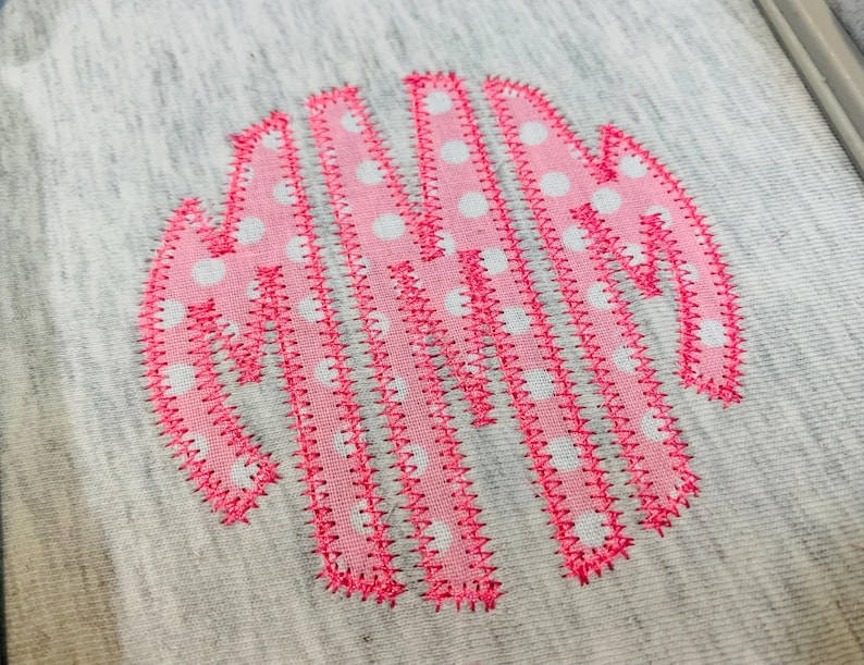 May include: A circular monogram design embroidered onto a heather grey fabric. The letters are formed from pink fabric featuring a white polka dot pattern, with the edges of each letter secured by a bright pink zigzag stitch embroidery thread.