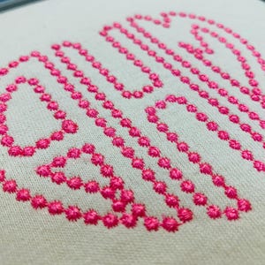 May include: A monogram design embroidered on off-white fabric using bright pink thread. The design features three stylised letters formed by a series of small, circular stitches, creating a textured, dotted outline effect on the light background.