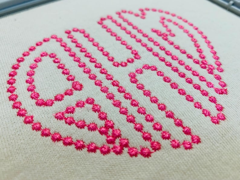 May include: A monogram design embroidered on off-white fabric using bright pink thread. The design features three stylised letters formed by a series of small, circular stitches, creating a textured, dotted outline effect on the light background.
