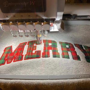 May include: An embroidery machine stitching the word "MERRY" onto a gray fabric. The letters are in a plaid pattern with red, green, and brown hues, outlined in red. The machine's needle is actively sewing the fabric, creating a festive holiday design.