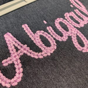 May include: Close-up of a dark gray fabric with the name "Abigail" embroidered in a light purple color. The embroidery has a textured, chain-like stitch. A white frame is visible at the top of the image.