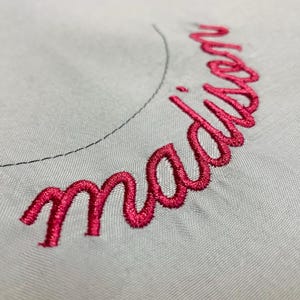 Op de afbeelding: Een close-up van het woord "madison" geborduurd in felroze op een lichtgrijze stof.