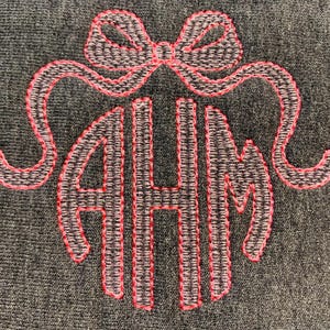 May include: Embroidered monogram with the letters AHM, encircled by a bow. The letters and bow are outlined in bright pink thread, set against a textured gray background. The design is likely for a personalized item.