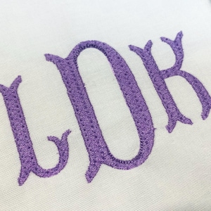 May include: A white fabric surface featuring the embroidered letters LDK in a purple thread. The letters are stitched in a decorative serif font with a visible texture created by the embroidery process, centered on the plain white background.