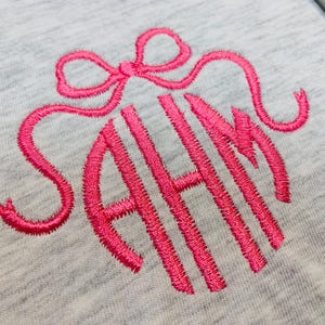 Op de afbeelding: Close-up van een lichtgrijze stof met een felroze geborduurd monogram. Het monogram toont de letters AHN in een gestileerd, afgerond lettertype, omcirkeld door een decoratieve strik.