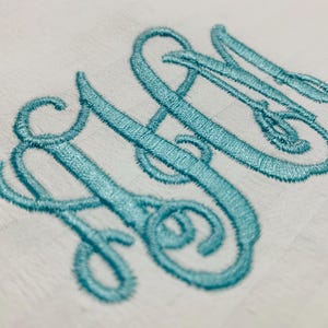 Op de afbeelding: Close-up van een witte stof met een lichtblauw geborduurd monogram. Het monogram toont de letters "SN" in een cursief, verweven ontwerp, met een mes- en vorkdetail.