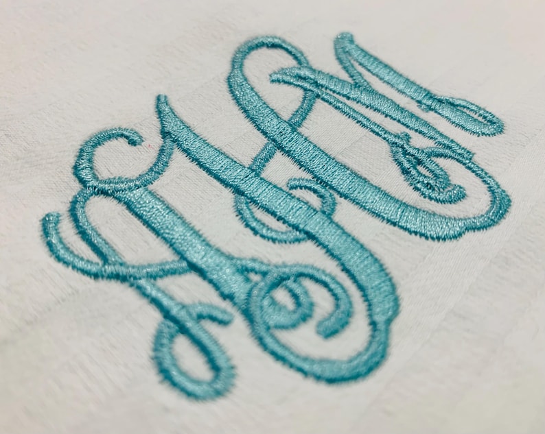 Op de afbeelding: Close-up van een witte stof met een lichtblauw geborduurd monogram. Het monogram toont de letters "SN" in een cursief, verweven ontwerp, met een mes- en vorkdetail.