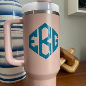 Puede incluir: Un vaso t&eacute;rmico rosa con asa y tapa blanca. El vaso presenta un dise&ntilde;o de monograma turquesa en forma hexagonal. El asa y la tapa tambi&eacute;n son rosas. Dise&ntilde;ado para mantener las bebidas calientes o fr&iacute;as.
