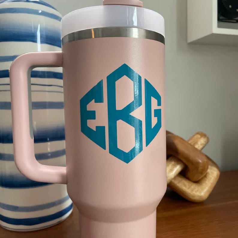 Puede incluir: Un vaso t&eacute;rmico rosa con asa y tapa blanca. El vaso presenta un dise&ntilde;o de monograma turquesa en forma hexagonal. El asa y la tapa tambi&eacute;n son rosas. Dise&ntilde;ado para mantener las bebidas calientes o fr&iacute;as.