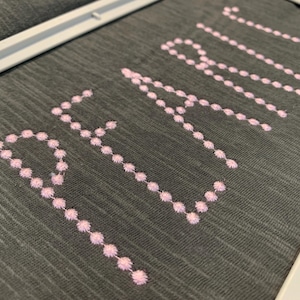 May include: A dark grey fabric surface framed in white, featuring the word PEARL embroidered in light pink thread. The letters are formed by a series of small, circular stitched dots, creating a textured and precise decorative design.