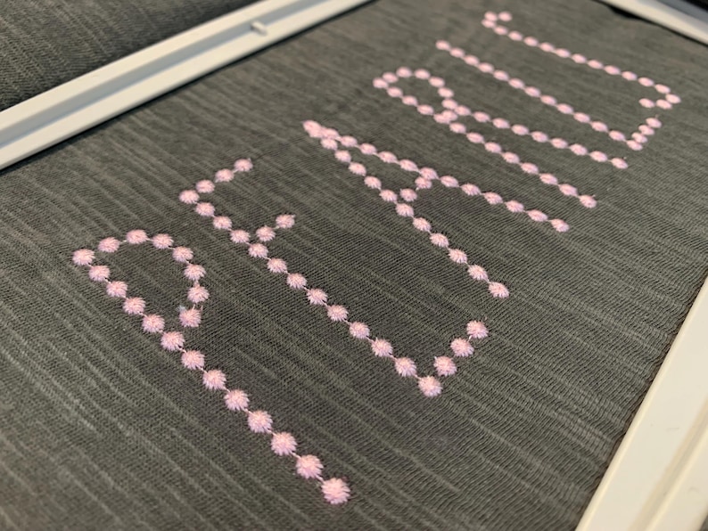 May include: A dark grey fabric surface framed in white, featuring the word PEARL embroidered in light pink thread. The letters are formed by a series of small, circular stitched dots, creating a textured and precise decorative design.