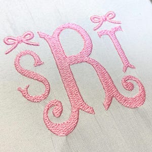 May include: A white fabric surface featuring pink machine embroidery of the letters S, R, and T in a cursive monogram style. The letters are accented with two small pink embroidered bows positioned above the letters S and T respectively.