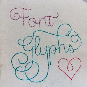 May include: A white fabric surface featuring embroidered text and a heart shape. The word Font is stitched in pink glitter thread, while the word Glyphs is stitched in teal glitter thread. A small heart is stitched in pink glitter thread below.