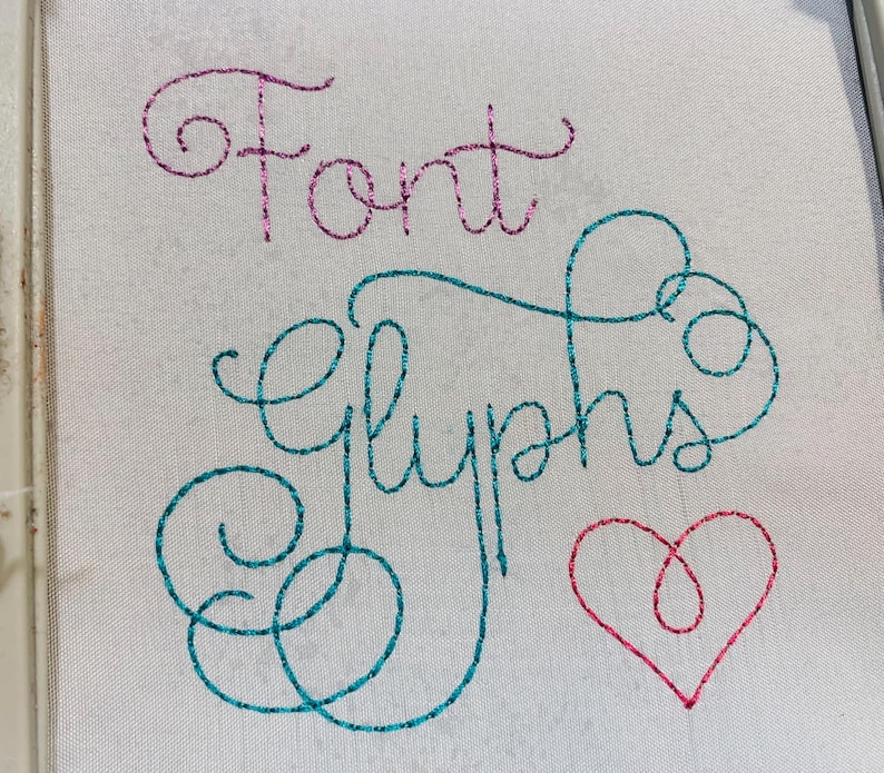May include: A white fabric surface featuring embroidered text and a heart shape. The word Font is stitched in pink glitter thread, while the word Glyphs is stitched in teal glitter thread. A small heart is stitched in pink glitter thread below.