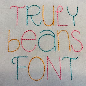 May include: A light gray fabric surface featuring the embroidered text "TRULY beans FONT" in a multi-colored stitch pattern. The letters are stitched in alternating shades of teal, orange, and pink, creating a vibrant and textured appearance.