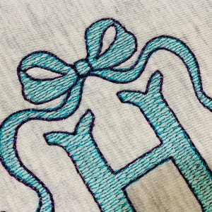 May include: Close-up of a light gray fabric with teal embroidery. The embroidery features a bow and the letter "H" with a purple outline. The stitching has a textured appearance.