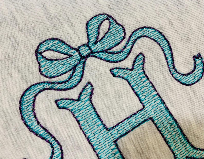May include: Close-up of a light gray fabric with teal embroidery. The embroidery features a bow and the letter "H" with a purple outline. The stitching has a textured appearance.