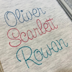 May include: A light gray heathered fabric featuring three lines of embroidered names in cursive script. The top name Oliver is stitched in blue, the middle name Scarlett is in red, and the bottom name Rowan is in teal thread.