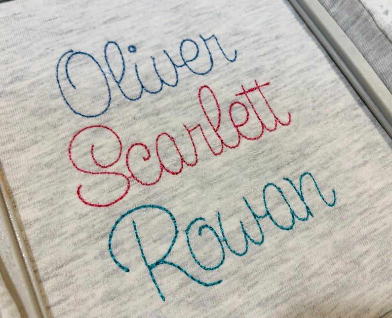 May include: A light gray heathered fabric featuring three lines of embroidered names in cursive script. The top name Oliver is stitched in blue, the middle name Scarlett is in red, and the bottom name Rowan is in teal thread.