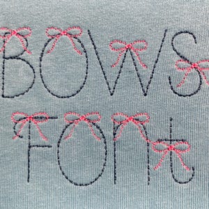 May include: A light blue fabric featuring dark navy blue embroidered lettering that spells out BOWS FONT. Each letter is accented with small, detailed pink embroidered bows placed at the top or sides of the characters in a decorative style.