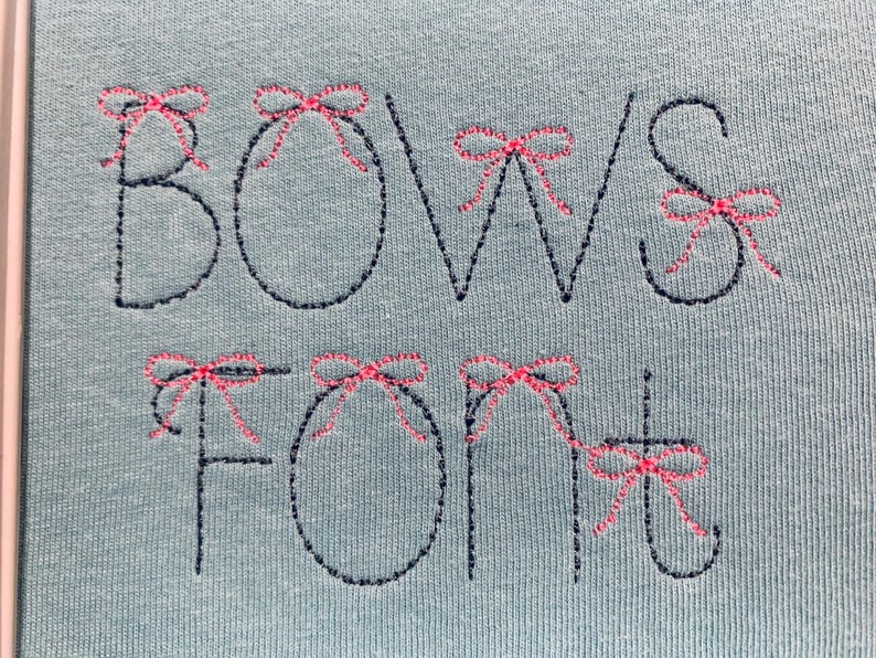 May include: A light blue fabric featuring dark navy blue embroidered lettering that spells out BOWS FONT. Each letter is accented with small, detailed pink embroidered bows placed at the top or sides of the characters in a decorative style.