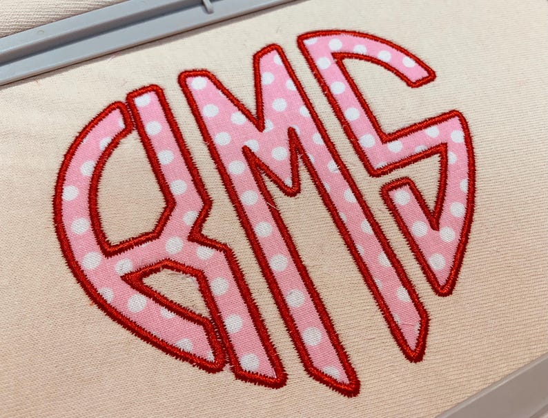 May include: An embroidered monogram featuring the letters RMS in a large, stylised font. The letters are light pink with white polka dots and are outlined in a bold, vibrant red thread. The design is stitched onto a plain, light beige fabric.