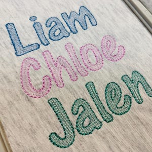 Puede incluir: Primer plano de una tela gris claro con los nombres bordados "Liam" en azul, "Chloe" en rosa y "Jalen" en verde. Los nombres están en una fuente juguetona y redondeada, cosidos con un contorno contrastante.