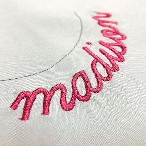 May include: A light gray fabric surface featuring the name Madison embroidered in a vibrant pink cursive script. A thin, curved black stitched line is visible above the embroidered text, indicating a circular embroidery placement guide.