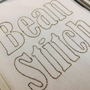 May include: Close-up of a white fabric with the words "Bean Stitch" embroidered in a gold-colored thread. The text is in a stylized, rounded font, and the stitching is visible.
