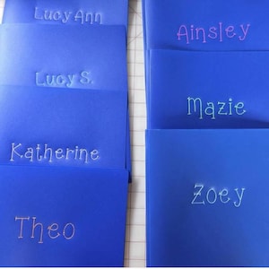 May include: A collection of bright blue rectangular folders, each featuring a name embroidered in a light-colored thread. The names visible on the folders are Lucy Ann, Lucy S., Katherine, Theo, Ainsley, Mazie, and Zoey, all in a simple font.