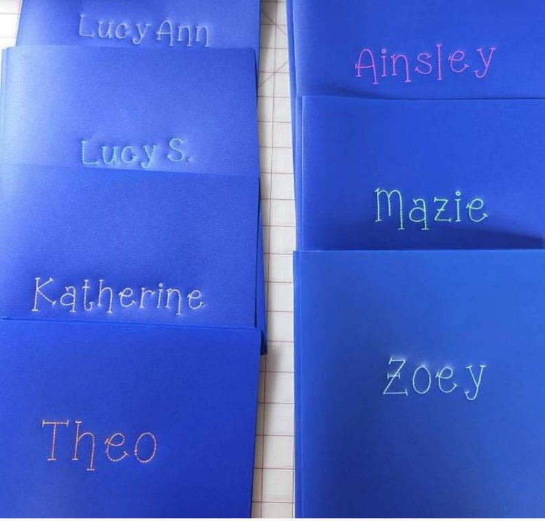 May include: A collection of bright blue rectangular folders, each featuring a name embroidered in a light-colored thread. The names visible on the folders are Lucy Ann, Lucy S., Katherine, Theo, Ainsley, Mazie, and Zoey, all in a simple font.