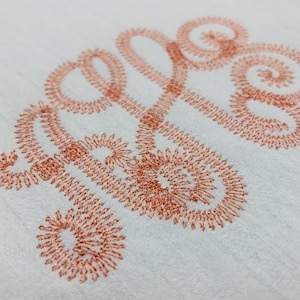 May include: A white fabric surface featuring an embroidered monogram in a metallic copper thread. The design consists of three intertwined cursive letters with intricate, swirling loops and a dense, textured stitch pattern throughout.