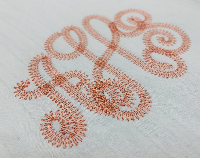 May include: A white fabric surface featuring an embroidered monogram in a metallic copper thread. The design consists of three intertwined cursive letters with intricate, swirling loops and a dense, textured stitch pattern throughout.