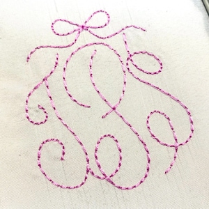 May include: A white fabric surface featuring a decorative monogram embroidered with bright pink thread. The design consists of elegant, flowing cursive lines that form a stylized letter, topped with a small, delicate bow detail in the same pink.