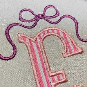 May include: Close-up of an embroidered design featuring a purple bow and the letter "F". The letter is pink with white stripes. The embroidery is on a light blue fabric.