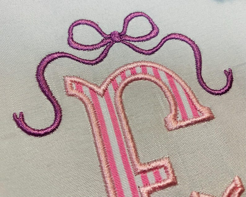 May include: Close-up of an embroidered design featuring a purple bow and the letter "F". The letter is pink with white stripes. The embroidery is on a light blue fabric.