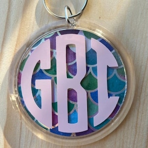 Puede incluir: Un llavero redondo con un borde de pl&aacute;stico transparente. El llavero presenta un monograma rosa con las letras GBI sobre un colorido patr&oacute;n de escamas de sirena en tonos de azul, verde y morado.