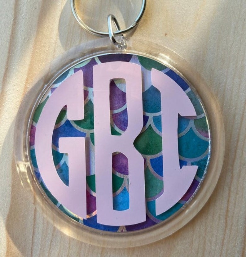 Puede incluir: Un llavero redondo con un borde de pl&aacute;stico transparente. El llavero presenta un monograma rosa con las letras GBI sobre un colorido patr&oacute;n de escamas de sirena en tonos de azul, verde y morado.