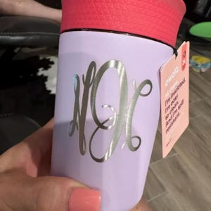 Puede incluir: Un vaso t&eacute;rmico lila con tapa roja. El vaso presenta un monograma plateado y una etiqueta que dice "Hi there, I'm insulated. I'm easy to clean. And I'm spill resistant."