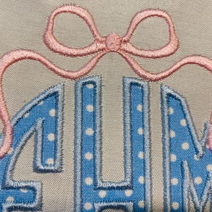 May include: Embroidered monogram in light blue with white polka dots, outlined in a darker blue. A pink bow is stitched above the letters. The background is a neutral beige fabric. The letters appear to be the initials of a name.