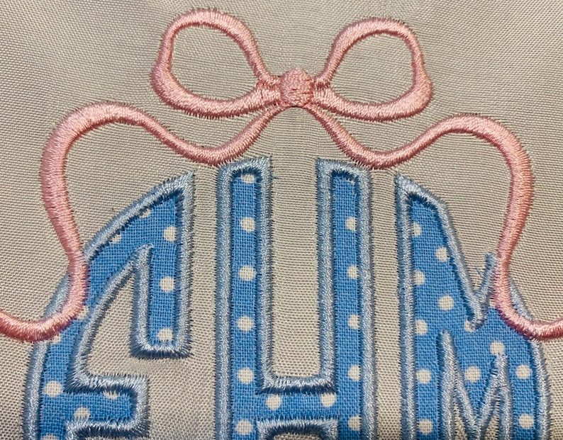 May include: Embroidered monogram in light blue with white polka dots, outlined in a darker blue. A pink bow is stitched above the letters. The background is a neutral beige fabric. The letters appear to be the initials of a name.