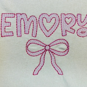 May include: Embroidered design on a cream-colored fabric. The name "EMORY" is stitched in pink thread, with a heart replacing the "O". A pink bow is embroidered below the name. The design is likely for a personalized item.