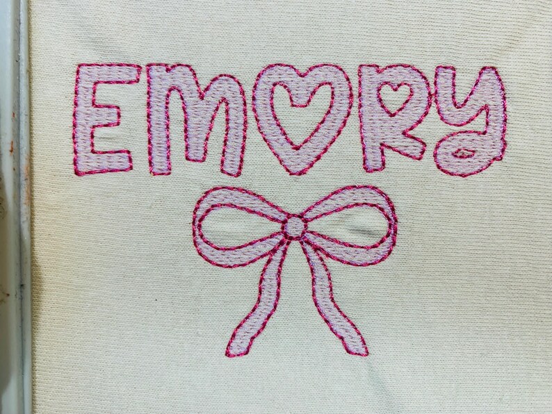 May include: Embroidered design on a cream-colored fabric. The name "EMORY" is stitched in pink thread, with a heart replacing the "O". A pink bow is embroidered below the name. The design is likely for a personalized item.