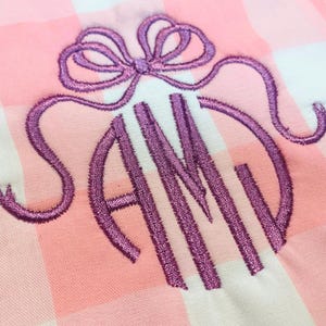 Op de afbeelding: Een lichtroze en witte gingham stof met een paars geborduurd monogram van de letters SAM, met een decoratieve strik direct boven de letters gestikt. Het borduurgaren heeft een lichte glans en een getextureerd uiterlijk.