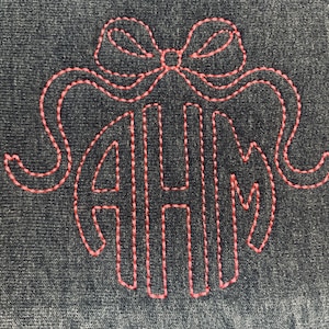 May include: A dark denim-colored fabric with a stitched design in coral thread. The design features a bow at the top and the letters AHM in a monogram style. The stitching is detailed and creates a textured effect.