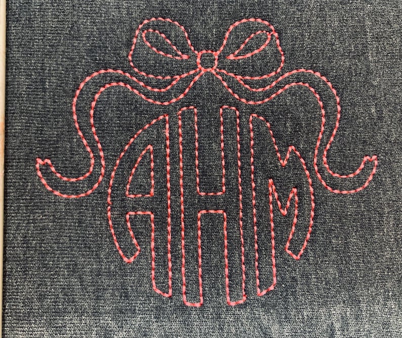 May include: A dark denim-colored fabric with a stitched design in coral thread. The design features a bow at the top and the letters AHM in a monogram style. The stitching is detailed and creates a textured effect.